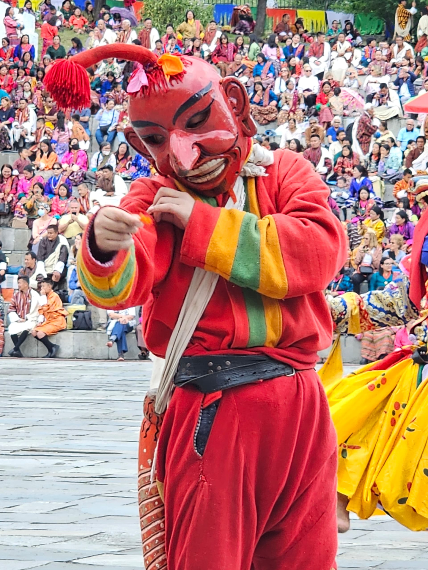 A red-masked festival performer in a bright robe before a crowd in traditional Bhutanese dress