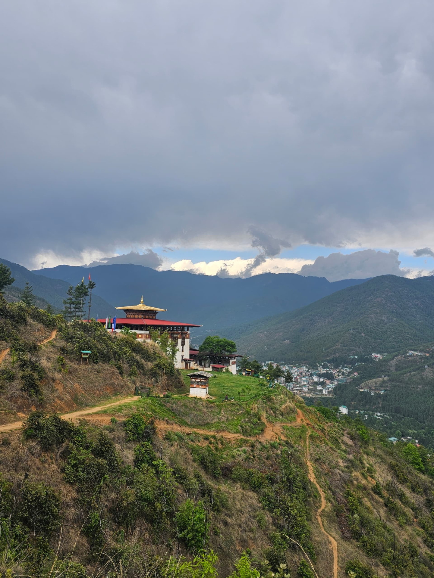 A whitewashed Bhutanese monastery with a tiered red roof and golden spire on a green hillside, overlooking a valley town beneath a storm-lit mountain sky